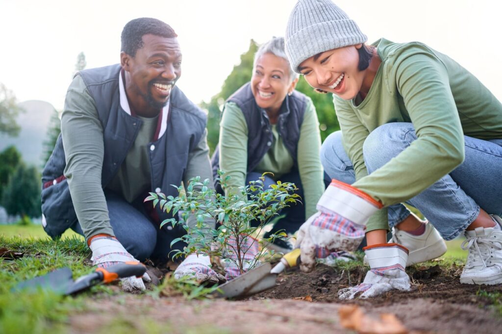 Three people smile as they kneel outdoors, planting a small shrub together with gardening tools and gloves, depicted in the image iStock-1479288162-min.