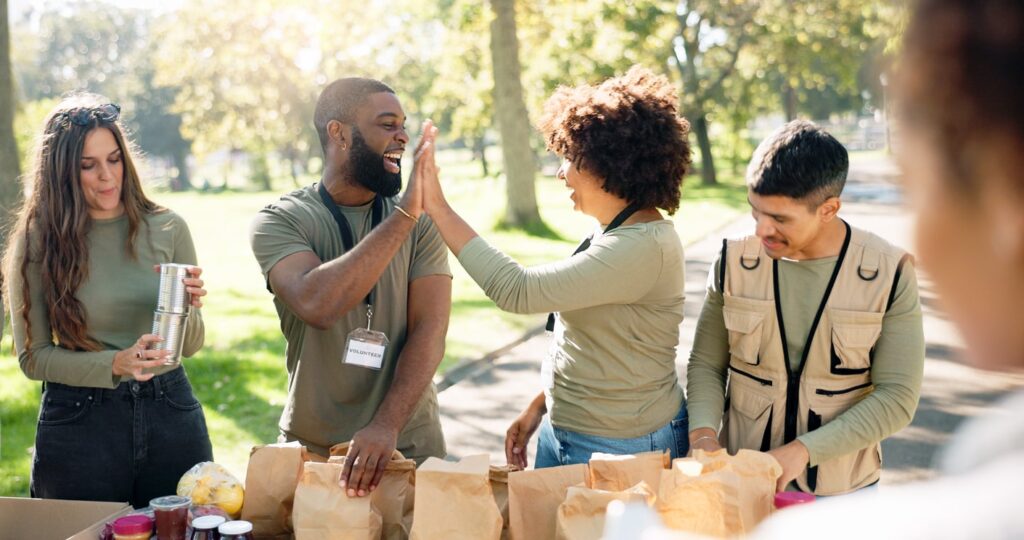 In iStock-2155998573-min, a group packs food items into paper bags at an outdoor table while two people in the center share a high-five.