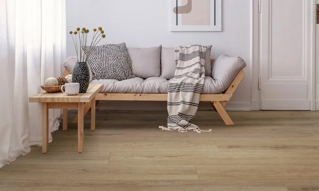 A minimalist living room with a wooden sofa, gray cushions, striped blanket, coffee table with a cup, and dominated by neutral decor features Room-Scene-DynamixGrande-Plank-Olive-(Single-width) light wood flooring.