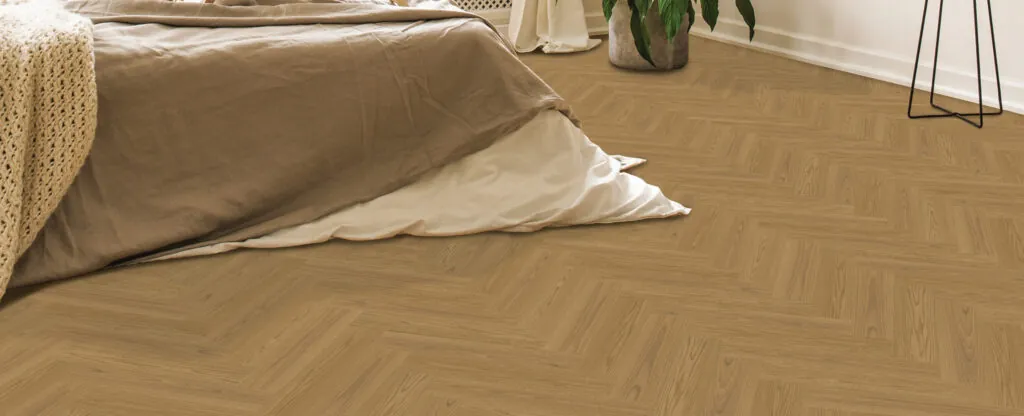 A bed with light brown bedding rests in a bedroom featuring Velvet Grove Herringbone-Radiant Ridge flooring, complemented by a potted plant in the corner.