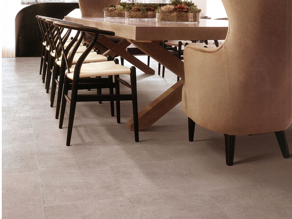 A modern dining room features a wooden table, eight black chairs with light cushions on one side, a beige upholstered chair at the end, all set atop Dynamix-Grande-Tile-Zirconia-web flooring.