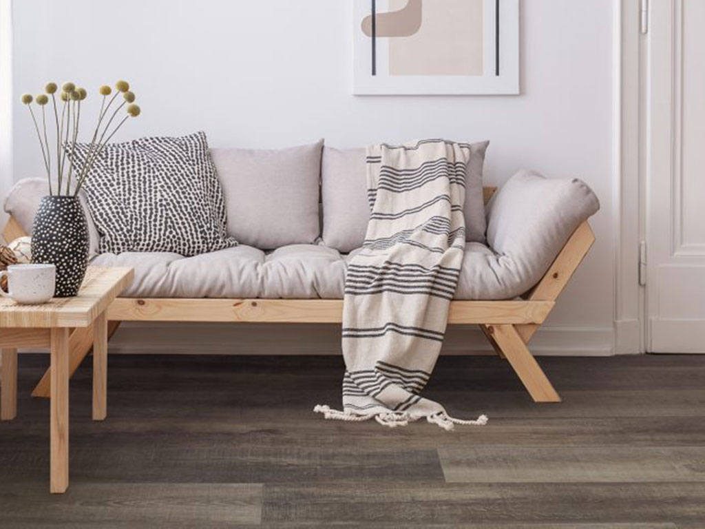 A minimalist living room features a wooden-framed sofa with neutral cushions, a striped throw, a small wooden table, and a vase with dried flowers on Room-Scene-Dynamix-Grande-Plank-Corinth flooring.