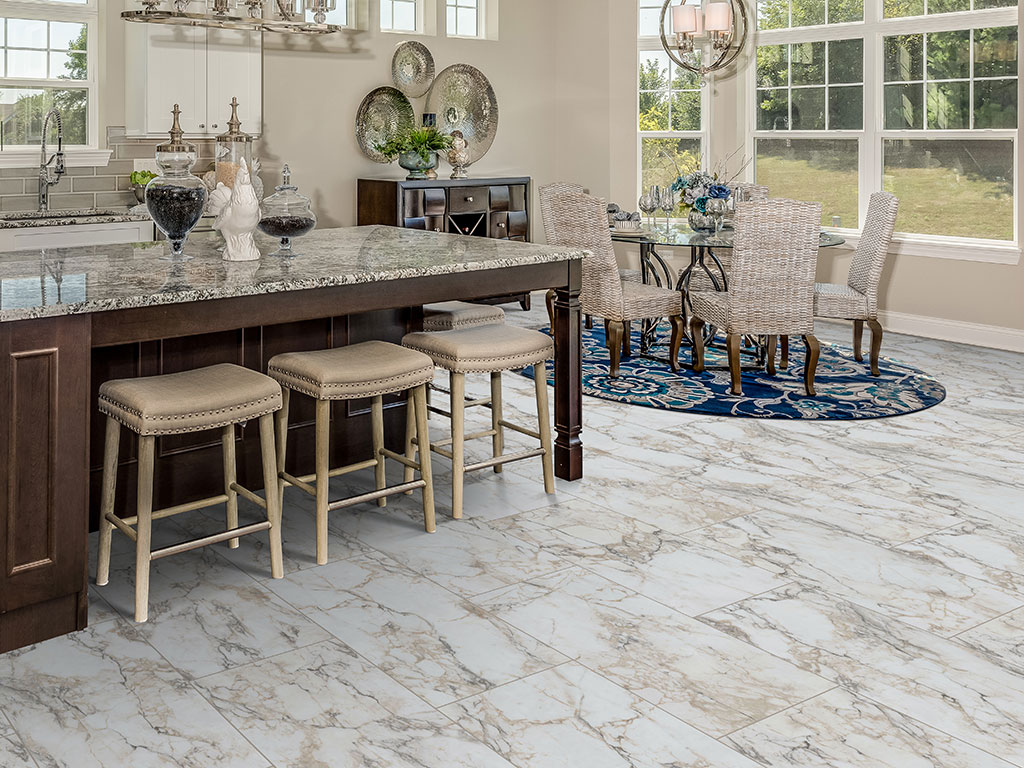 Kitchen featuring Room-Scene-Dynamix-XL-Tile-Blanc-web flooring, a granite island with three upholstered stools, a dining table with wicker chairs, blue patterned rug, and large windows for natural light.